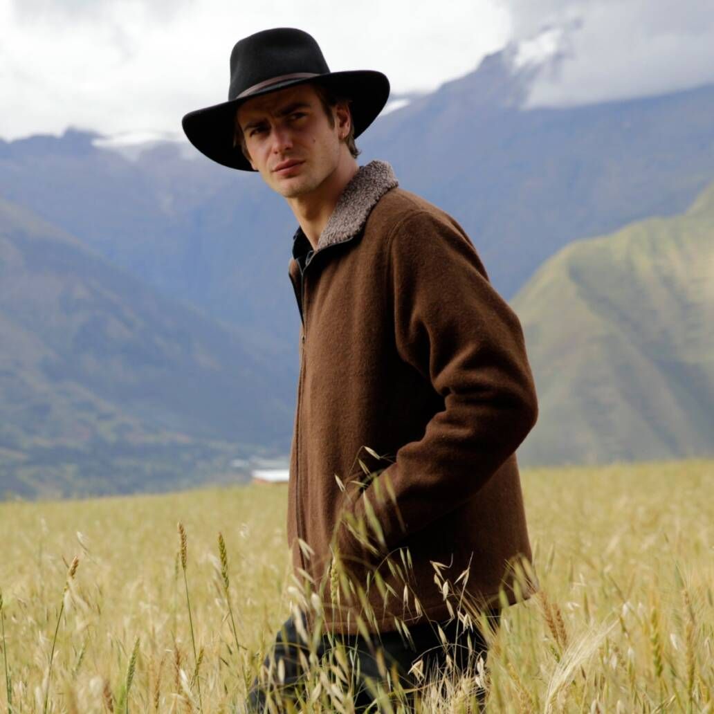 A young man wearing a brimmed hat with his hands in the pockets displays the alpaca barn jacket in a prairie scene with grass to the hips and mountains in the background