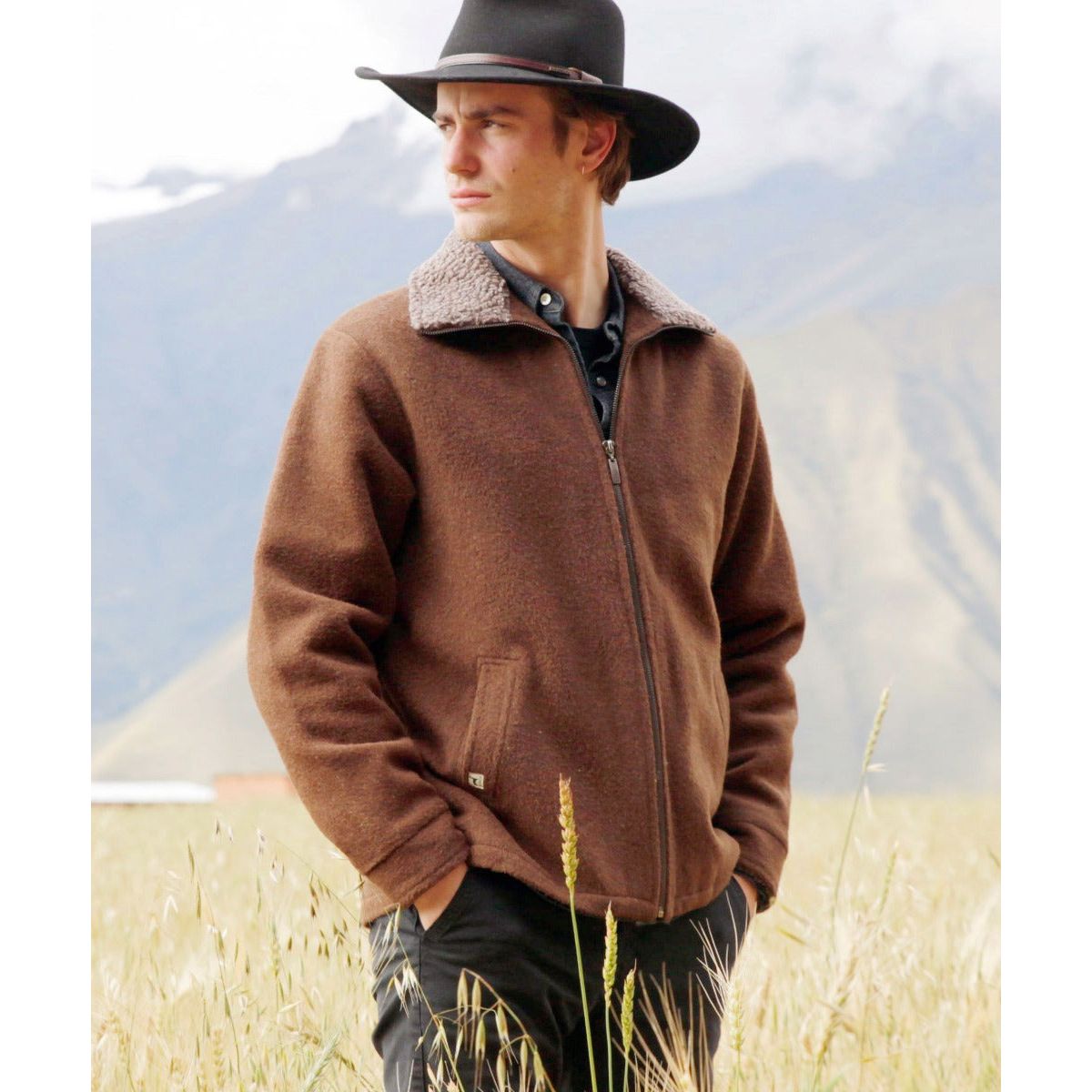 young man wearing brown alpaca jacket and brimmed hat standing facing right but looking left in dramatic fashion